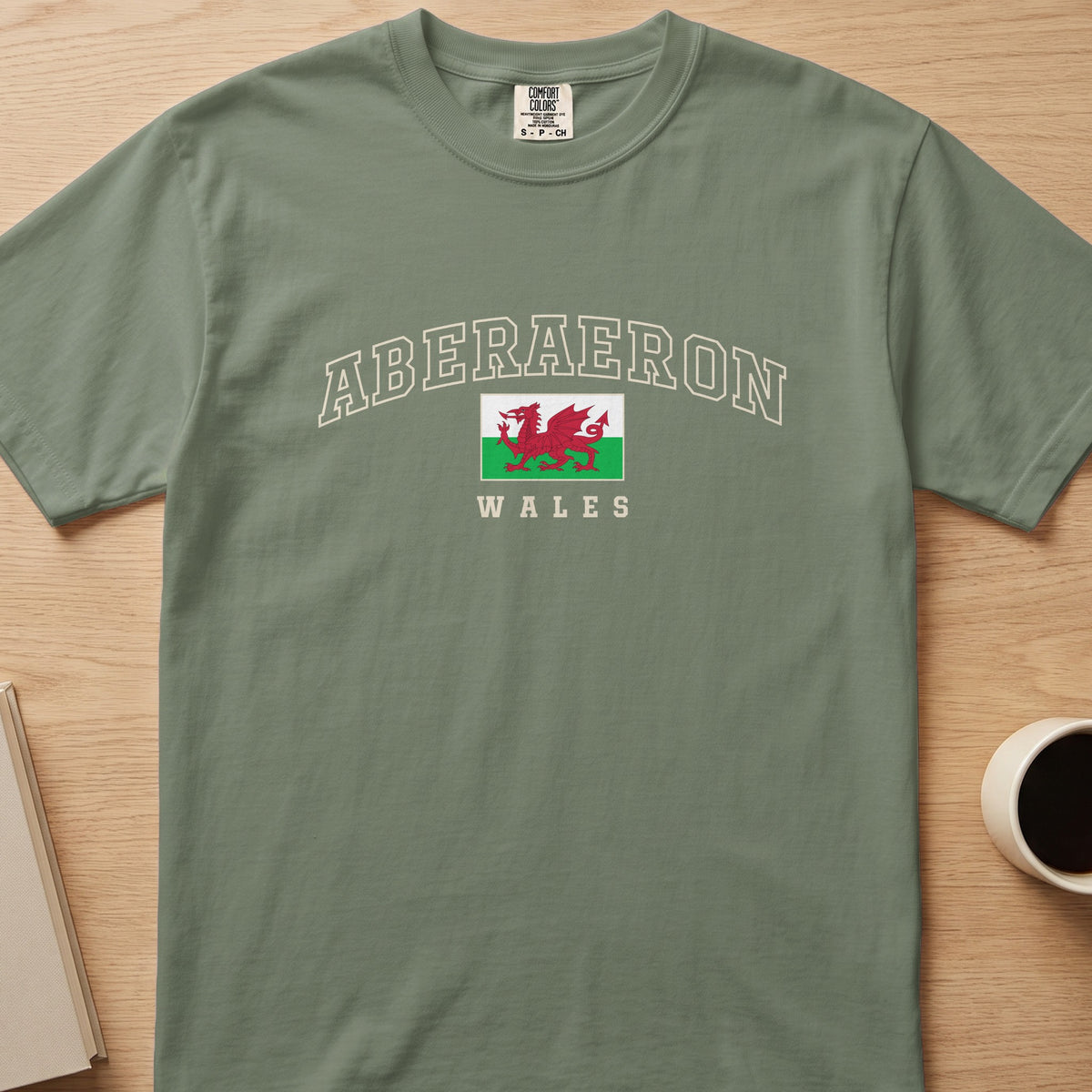 Moss green Aberaeron Wales T-shirt laid flat, with cream collegiate outline lettering and a red Welsh dragon flag.