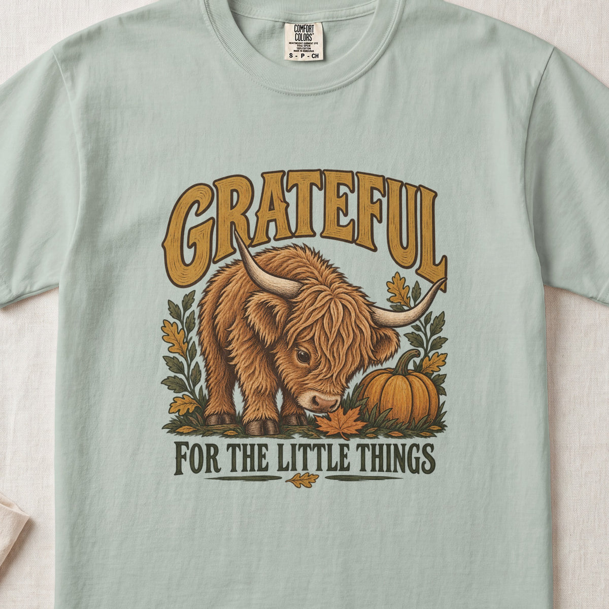 Grateful for the Little Things Highland cow design on a Bay Comfort Colors 1717 T-shirt, flat lay