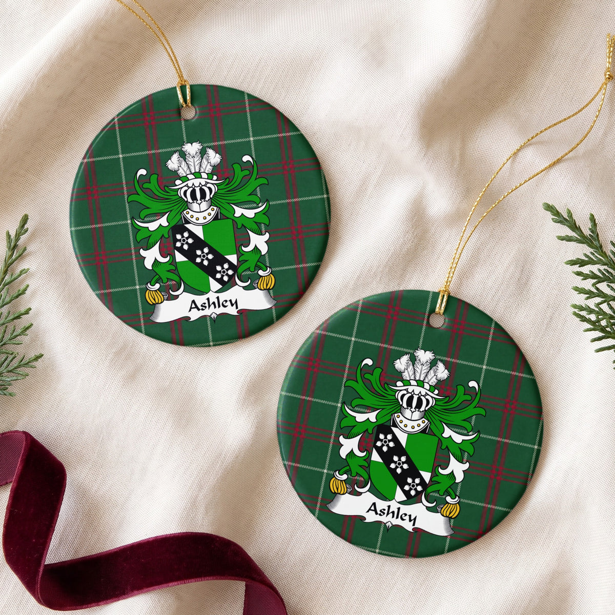 Two Ashley ornaments on cream fabric with evergreen and burgundy ribbon; a green-and-white shield crossed by a black band with three white flower-like motifs on Welsh national tartan.