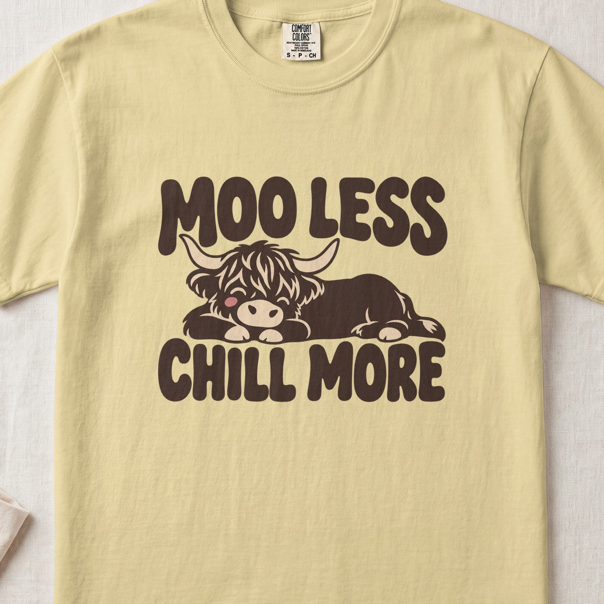 Moo Less, Chill More Highland cow design on a Butter Comfort Colors 1717 T-shirt, flat lay