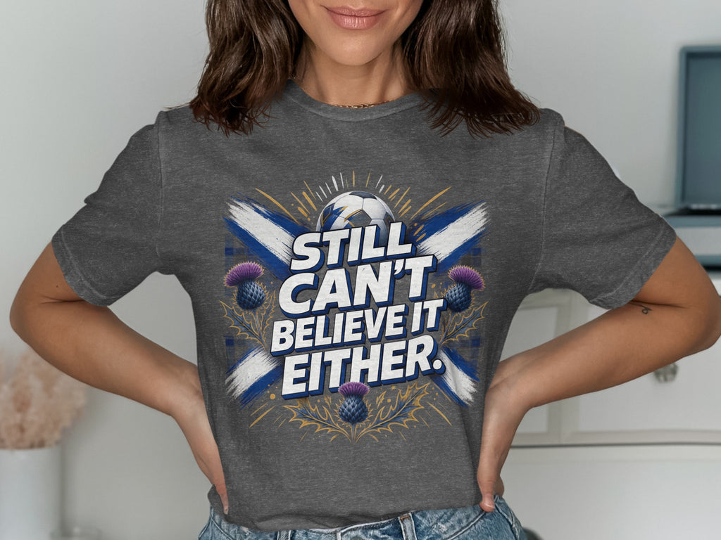 Dark grey t-shirt with Scotland flag, thistle, and text "STILL CAN'T BELIEVE IT EITHER."