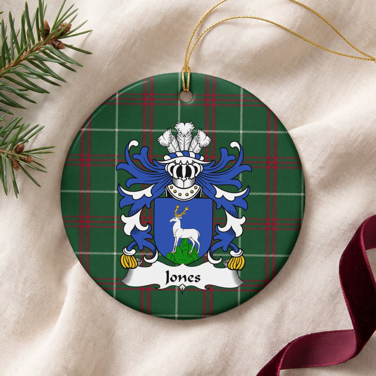 Jones Style 1 ornament on cream fabric with gold hanging string, evergreen and burgundy ribbon; a blue shield with a white stag standing on a green mound on Welsh national tartan.