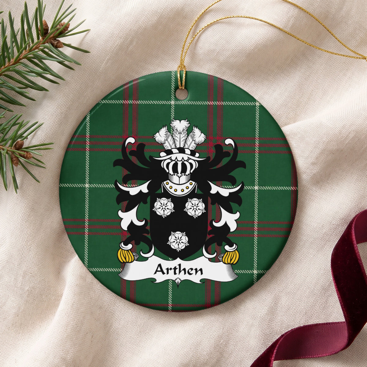 Arthen family crest and surname on a round Welsh national tartan ornament with a gold cord, resting on cream fabric beside an evergreen sprig and burgundy ribbon.