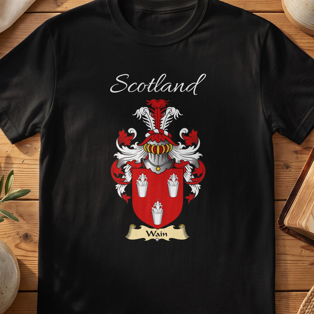 Black Wain Scottish family crest T-shirt laid flat, showing “Scotland” above a red-and-silver shield with three pale bird-like figures.