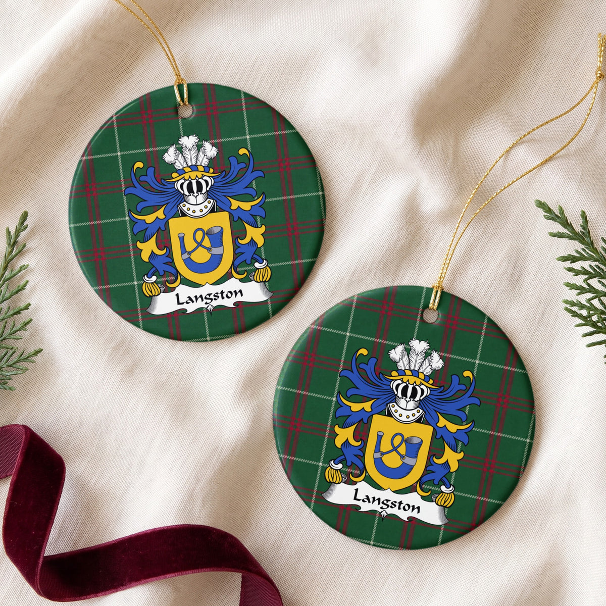 Two Langston ornaments on cream fabric beside evergreen sprigs and burgundy ribbon, showing a gold shield with a blue hunting horn and looped cord on Welsh national tartan.