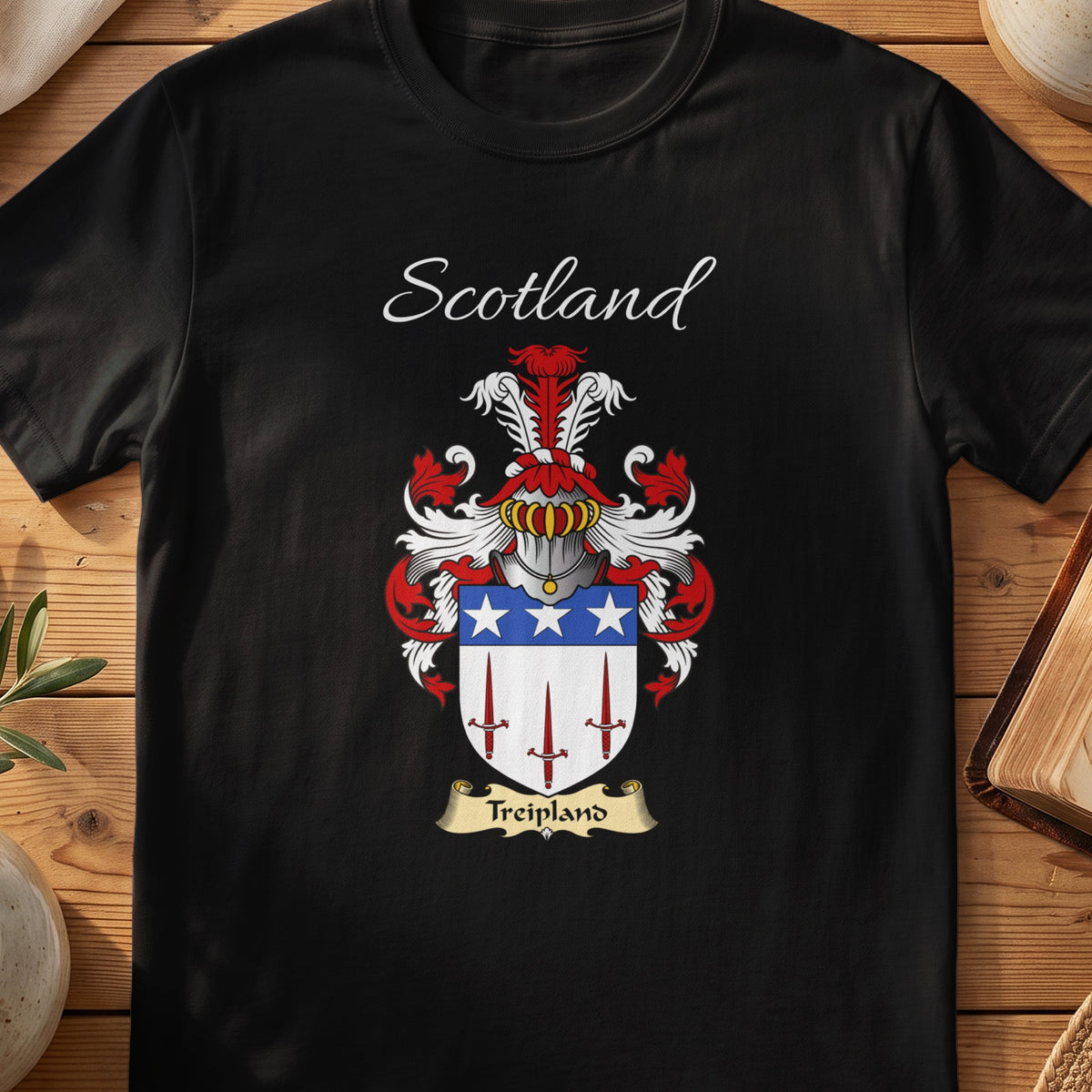 Black Treipland Scottish family crest T-shirt laid flat, showing “Scotland” above a red-blue-and-silver shield with stars and slender vertical devices.
