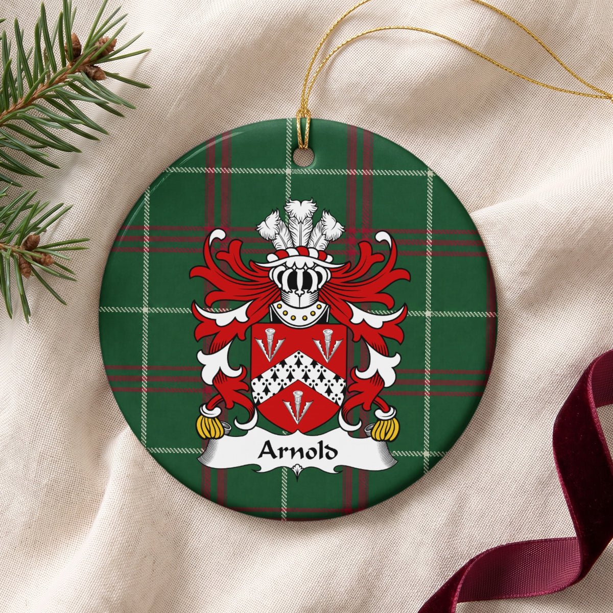 Arnold family crest and surname on a round Welsh national tartan ornament with a gold cord, resting on cream fabric beside an evergreen sprig and burgundy ribbon.