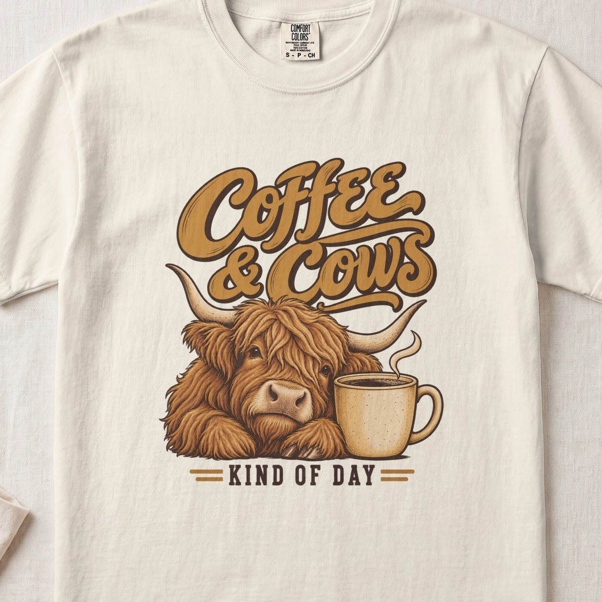 Coffee & Cows Kind of Day Highland cow design on an Ivory Comfort Colors 1717 T-shirt, flat lay