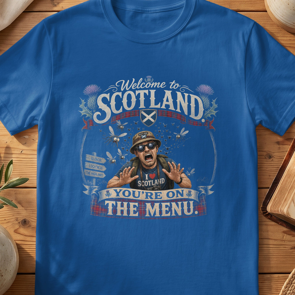 Blue t-shirt graphic: Man with scared expression surrounded by flying insects, with "Welcome to Scotland" and "You're on the Menu." text.