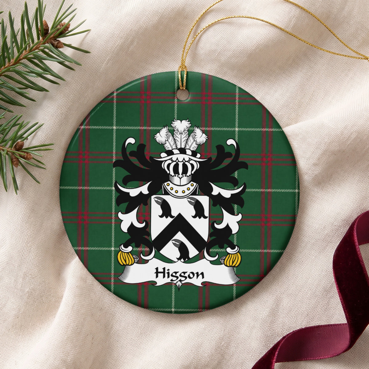 Higgon ornament on cream fabric beside evergreen and burgundy ribbon, with gold string; three black bird heads and a black chevron on a white shield.