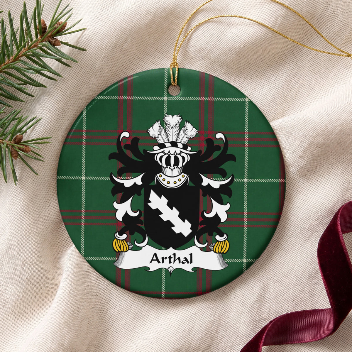 Arthal family crest and surname on a round Welsh national tartan ornament with a gold cord, resting on cream fabric beside an evergreen sprig and burgundy ribbon.